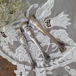 May include: Three silver-toned metal sugar tongs with ornate embossed handles and claw-shaped ends. Each tong features a distinct decorative pattern, including floral motifs and shield-like shapes, with a polished metallic finish throughout.