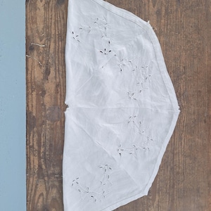 Puede incluir: Una pieza de tela blanca en forma de semic&iacute;rculo con un delicado bordado de ojetes en un patr&oacute;n de enredadera floral. Los bordes est&aacute;n rematados con una costura fina y el tejido presenta un aspecto ligero y semitransparente con calados.