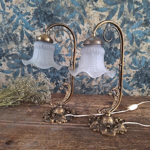 Puede incluir: Dos lámparas de mesa antiguas de latón con pantallas de vidrio esmerilado. Las lámparas tienen un diseño floral en la base y un brazo curvo. Las pantallas en forma de campana tienen un patrón texturizado. Las lámparas están sobre una superficie de madera.