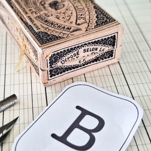 K&ouml;nnte beinhalten: Eine rechteckige Vintage-Schachtel mit verzierter schwarzer Typografie und dekorativen Mustern auf hellrosa Hintergrund, mit dem Text Volume Gauloise, Summerville & Co Birmingham und D&eacute;pos&eacute; Selon La, neben einer wei&szlig;en Karte mit einem schwarzen B.