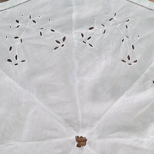 Puede incluir: Una pieza textil circular blanca con delicados bordados calados en un patr&oacute;n de enredaderas florales. La tela presenta una abertura central y una costura decorativa en el borde exterior, mostrando un dise&ntilde;o claro con un trabajo de aguja detallado.