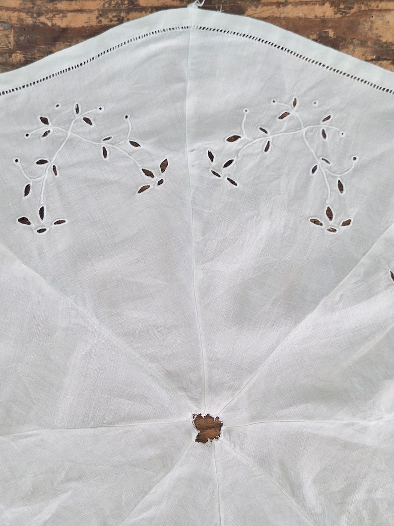Puede incluir: Una pieza textil circular blanca con delicados bordados calados en un patr&oacute;n de enredaderas florales. La tela presenta una abertura central y una costura decorativa en el borde exterior, mostrando un dise&ntilde;o claro con un trabajo de aguja detallado.