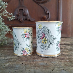 Könnte beinhalten: Zwei weiße Keramikbehälter mit floralen Mustern in Rosa, Gelb und Grün. Die Behälter haben goldfarbene Verzierungen. Sie stehen auf einer Holzoberfläche, mit einer dunklen geschnitzten Holztafel im Hintergrund.