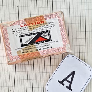 K&ouml;nnte beinhalten: Eine rechteckige Pappschachtel f&uuml;r A. Sommerville & Co. Stifte mit roten Zierr&auml;ndern und einem zentralen Etikett mit dem Text CAUTION, MARQUE DEPOSEE und MADE IN GREAT BRITAIN. Daneben liegt ein wei&szlig;es Papieretikett mit einem schwarzen Buchstaben A.