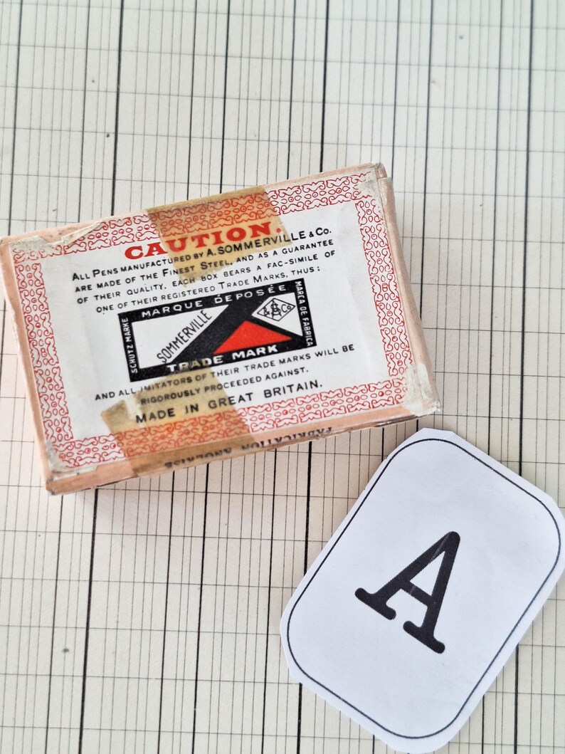 K&ouml;nnte beinhalten: Eine rechteckige Pappschachtel f&uuml;r A. Sommerville & Co. Stifte mit roten Zierr&auml;ndern und einem zentralen Etikett mit dem Text CAUTION, MARQUE DEPOSEE und MADE IN GREAT BRITAIN. Daneben liegt ein wei&szlig;es Papieretikett mit einem schwarzen Buchstaben A.