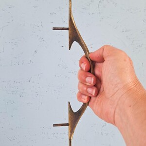 May include: A long, curved, antique-style brass door handle with a textured, weathered finish. The handle features two protruding threaded metal bolts on the inner side for mounting purposes, designed for attachment to a door or cabinet surface.