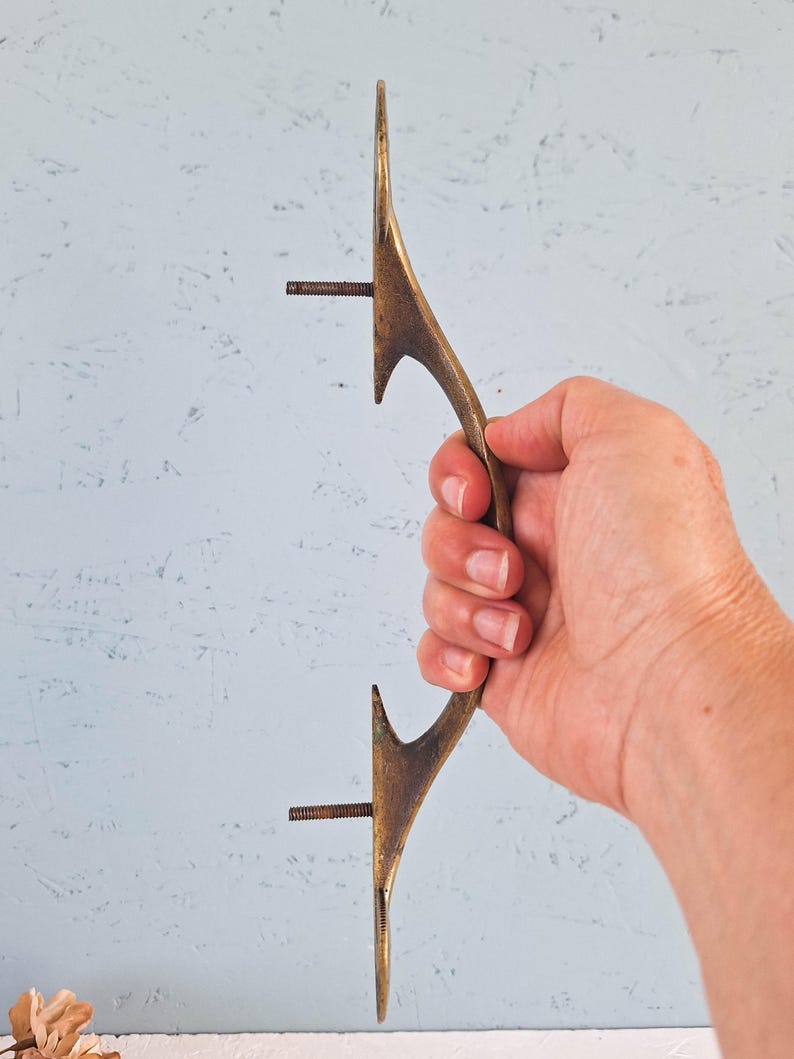 May include: A long, curved, antique-style brass door handle with a textured, weathered finish. The handle features two protruding threaded metal bolts on the inner side for mounting purposes, designed for attachment to a door or cabinet surface.