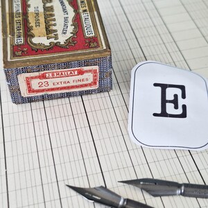 K&ouml;nnte beinhalten: Eine rechteckige Pappschachtel mit der Aufschrift J.B. MALLAT COMMANDANT BARATIER 100 PLUMES METALLIQUES N-23 POINTES EXTRA-FINES, ein wei&szlig;er Papierausschnitt mit einem schwarzen Buchstaben E und zwei Metallfedern auf linierten Papier.