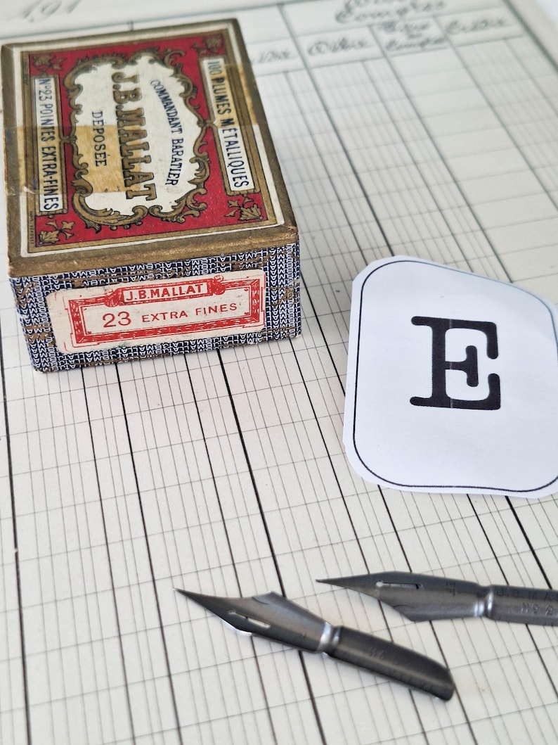 K&ouml;nnte beinhalten: Eine rechteckige Pappschachtel mit der Aufschrift J.B. MALLAT COMMANDANT BARATIER 100 PLUMES METALLIQUES N-23 POINTES EXTRA-FINES, ein wei&szlig;er Papierausschnitt mit einem schwarzen Buchstaben E und zwei Metallfedern auf linierten Papier.