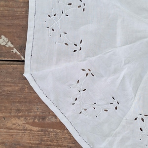 Puede incluir: Un tejido blanco con delicados bordados de ojal en un patr&oacute;n repetitivo de enredaderas florales. Los bordes est&aacute;n rematados con un dobladillo recto y un detalle de punto de escalera, mostrando una textura blanca limpia y n&iacute;tida.
