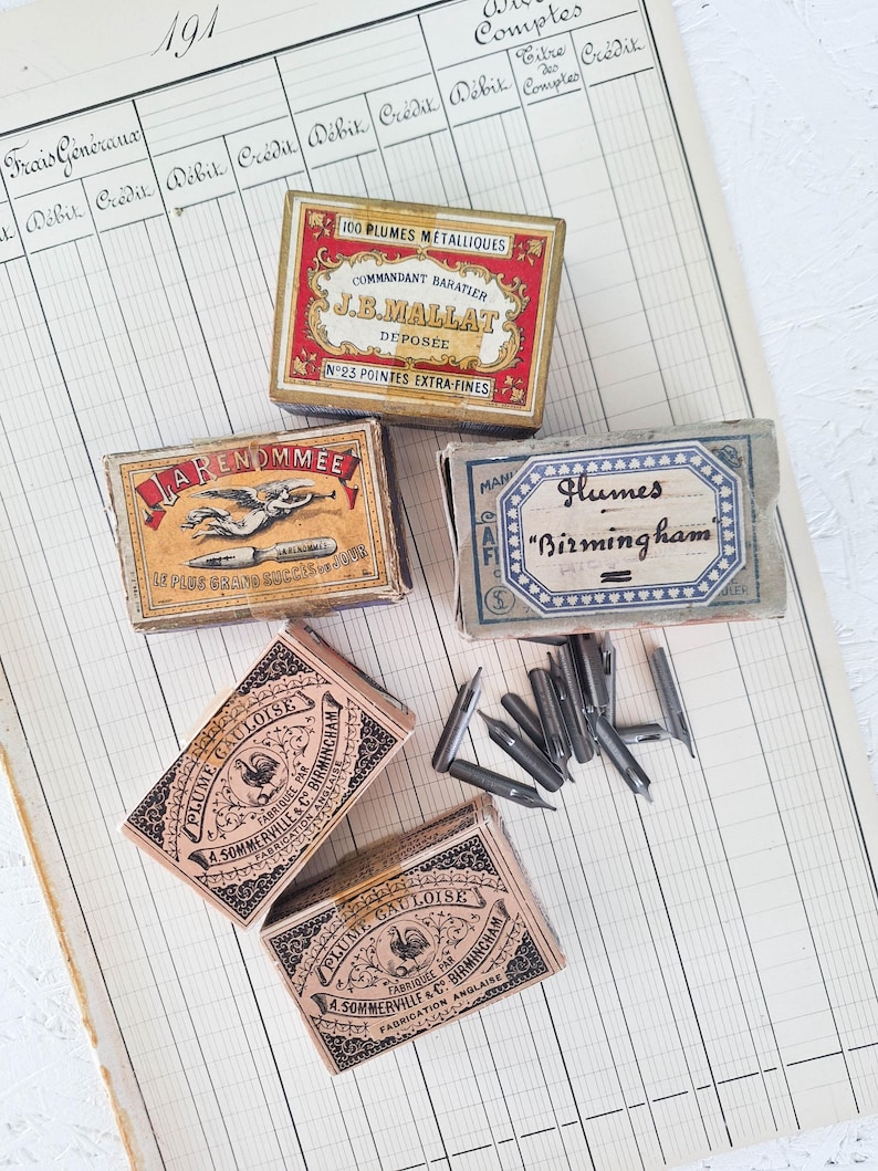 K&ouml;nnte beinhalten: F&uuml;nf rechteckige Pappschachteln mit metallenen Schreibfedern. Die Schachteln zeigen historische Typografie und Illustrationen, darunter Aufschriften wie J.B. Mallat, La Renommee und Plume Gauloise. Mehrere lose silberfarbene Schreibfedern liegen daneben.