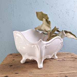 Puede incluir: Una jardinera de cerámica blanca con un diseño ornamentado. La jardinera tiene forma rectangular con bordes curvos y se apoya en cuatro patas pequeñas. Se colocan hojas y tallos secos dentro de la jardinera. El fondo es una pared azul claro.