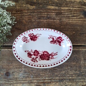Puede incluir: Una fuente de servir ovalada de cerámica blanca con un patrón floral de color rojo oscuro de claveles en el centro y un borde decorativo de pequeños triángulos rojos a lo largo del borde, ofreciendo una pieza funcional y limpia.