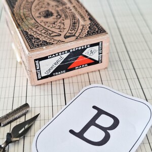 K&ouml;nnte beinhalten: Eine rechteckige Vintage-Pappschachtel mit der Aufschrift Plume G. Louise A. Sommerville & Co. in Schwarz und Pfirsich, eine Metall-Kalligrafiefeder und eine wei&szlig;e Papierkarte mit einem gro&szlig;en schwarzen Buchstaben B in Serifenschrift.
