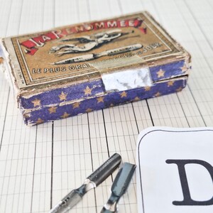 K&ouml;nnte beinhalten: Eine rechteckige Pappschachtel mit der Aufschrift LA RENOMMEE, einer Grafik einer gefl&uuml;gelten Figur und einer Feder, mit einer blauen Seite mit goldenen Sternen. Zwei metallische Kalligrafiefedern liegen vor der Schachtel neben einer wei&szlig;en Karte mit dem Buchstaben D.