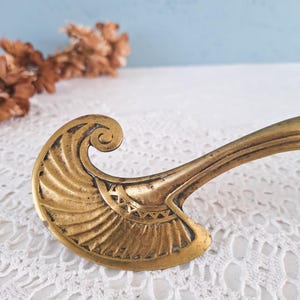 May include: A brass furniture handle featuring a curved, fan-like end with engraved radiating lines and a small triangular pattern near the base. The metal has a warm, aged patina with darker recessed areas highlighting the intricate design details.