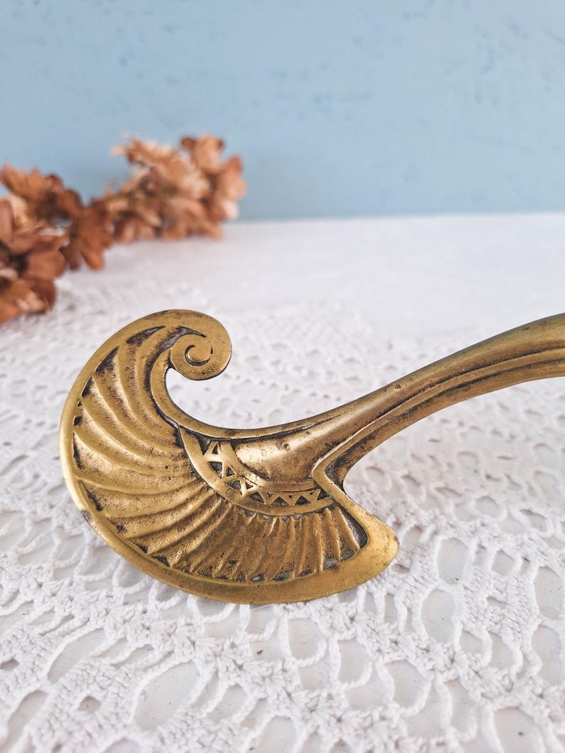 May include: A brass furniture handle featuring a curved, fan-like end with engraved radiating lines and a small triangular pattern near the base. The metal has a warm, aged patina with darker recessed areas highlighting the intricate design details.