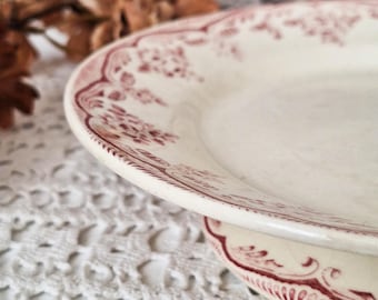 Puede incluir: Un plato de cerámica de color crema con un patrón decorativo floral y de volutas de color granate a lo largo del borde. El plato tiene un borde ligeramente elevado y un acabado liso y brillante, mostrando detalles botánicos.