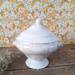 May include: A white ceramic tureen with a lid, handles, and a pedestal base. The tureen has a decorative finial on the lid. The exterior has a speckled, aged appearance. The background is a floral pattern on a yellow and brown surface.