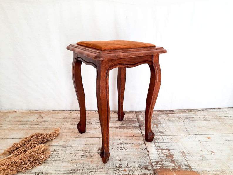 Peut inclure: Un petit tabouret en bois antique avec une assise carr&eacute;e et rembourr&eacute;e. Le tabouret a quatre pieds incurv&eacute;s et est en bois brun fonc&eacute;. Le coussin de si&egrave;ge est de couleur brun-orang&eacute; chaud. Le tabouret mesure environ 46 cm de haut.
