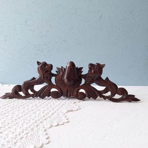 Puede incluir: Una pieza decorativa de madera tallada de color marrón oscuro con dos figuras de león que flanquean un diseño floral central. La pieza tiene detalles intrincados y una base de volutas, adecuada para la decoración de paredes.