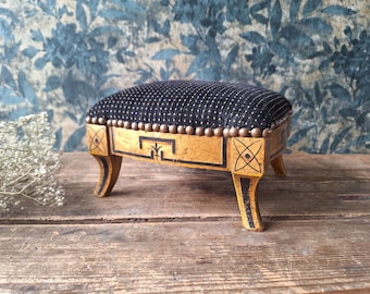 May include: A small footstool with a black fabric top featuring white pin-striping and a row of bronze-toned nailhead trim. The wooden base is painted in a distressed gold finish with black geometric line accents and four curved legs.