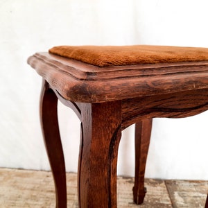 Peut inclure: Petit tabouret en bois de couleur marron fonc&eacute;. Il pr&eacute;sente des pieds courb&eacute;s et une assise carr&eacute;e recouverte d'un tissu de couleur rouille. Le grain du bois est visible, ajoutant &agrave; son charme vintage. Hauteur d'environ 40 cm.
