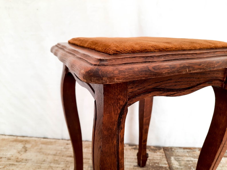 Peut inclure: Petit tabouret en bois de couleur marron fonc&eacute;. Il pr&eacute;sente des pieds courb&eacute;s et une assise carr&eacute;e recouverte d'un tissu de couleur rouille. Le grain du bois est visible, ajoutant &agrave; son charme vintage. Hauteur d'environ 40 cm.