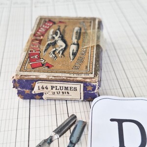 K&ouml;nnte beinhalten: Eine rechteckige Vintage-Pappschachtel mit beigem Deckel, der eine schwarze Illustration eines Engels und einer Schreibfeder zeigt, beschriftet mit "J. ARENMMET" und "LE PLUS GRAND". Die Seiten sind violett mit goldenen Sternen und "144 PLUMES" bedruckt. Zwei Metallfedern liegen daneben.