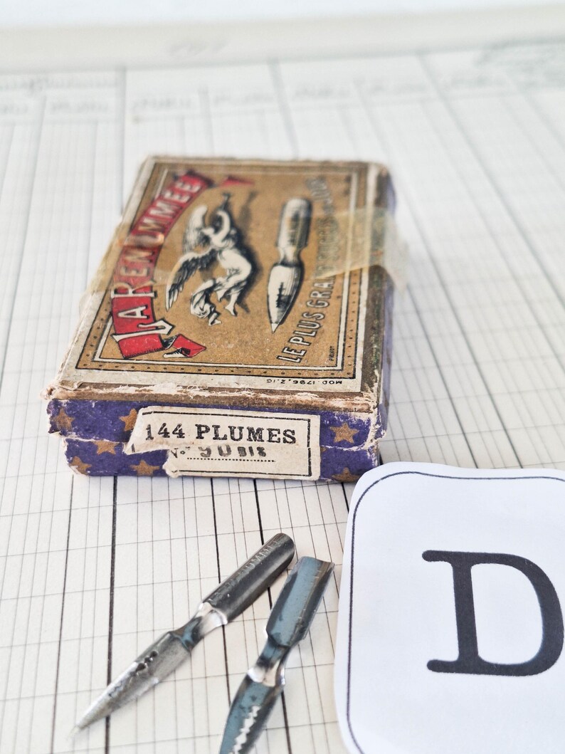 K&ouml;nnte beinhalten: Eine rechteckige Vintage-Pappschachtel mit beigem Deckel, der eine schwarze Illustration eines Engels und einer Schreibfeder zeigt, beschriftet mit "J. ARENMMET" und "LE PLUS GRAND". Die Seiten sind violett mit goldenen Sternen und "144 PLUMES" bedruckt. Zwei Metallfedern liegen daneben.