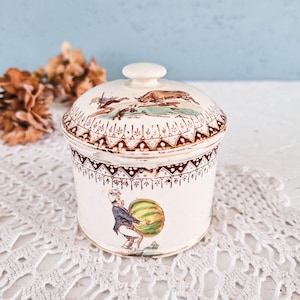 May include: A cream-colored ceramic lidded jar featuring a brown geometric border pattern. The lid depicts a hunting scene with a dog and a figure, while the body shows a figure in a blue coat and white hat carrying a large green striped melon.