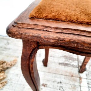 Peut inclure: Gros plan sur un tabouret en bois vintage avec des pieds incurv&eacute;s. Le tabouret a une finition marron fonc&eacute; vieillie avec une assise rectangulaire recouverte d'un tissu marron textur&eacute;. Le grain du bois est visible, ajoutant &agrave; son charme antique.