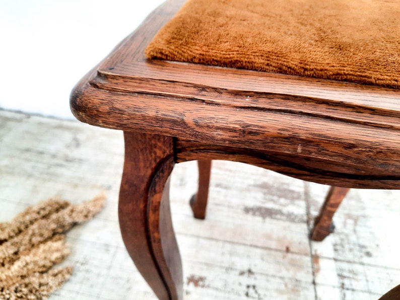 Peut inclure: Gros plan sur un tabouret en bois vintage avec des pieds incurv&eacute;s. Le tabouret a une finition marron fonc&eacute; vieillie avec une assise rectangulaire recouverte d'un tissu marron textur&eacute;. Le grain du bois est visible, ajoutant &agrave; son charme antique.