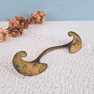 May include: A brass drawer pull with a curved, arching handle and two wide, scroll-shaped mounting plates at each end. The metal surface shows a weathered, aged patina with dark spots and a dull gold finish, featuring two small metal pins.