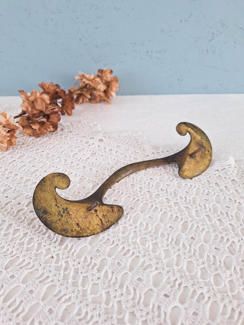 May include: A brass drawer pull with a curved, arching handle and two wide, scroll-shaped mounting plates at each end. The metal surface shows a weathered, aged patina with dark spots and a dull gold finish, featuring two small metal pins.