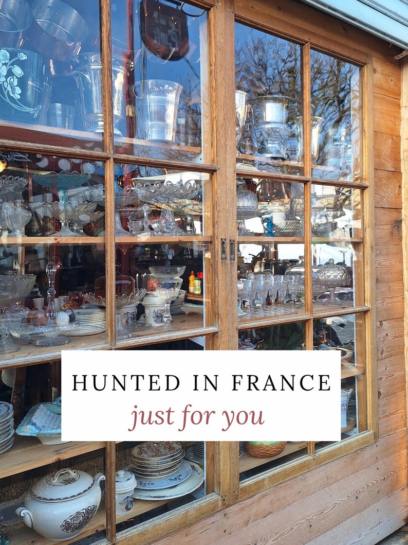 Peut inclure: Une vitrine en bois remplie de divers objets anciens en verre et en porcelaine. Le texte "HUNTED IN FRANCE just for you" est affich&eacute; sur un rectangle blanc. La fen&ecirc;tre refl&egrave;te l'environnement ext&eacute;rieur.