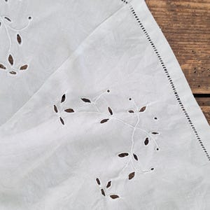 Puede incluir: Un tejido blanco con delicados bordados de ojal en un patr&oacute;n repetitivo de hojas y enredaderas. Los bordes est&aacute;n acabados con un detalle de dobladillo preciso, mostrando una superficie blanca limpia y n&iacute;tida con intrincados motivos florales.