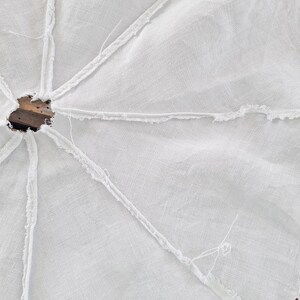 Puede incluir: Una falda de &aacute;rbol de tela blanca de forma circular con una abertura central y costuras radiales con bordes deshilachados. El tejido tiene una textura visible y est&aacute; construido con m&uacute;ltiples paneles unidos mediante costuras expuestas y sin rematar.