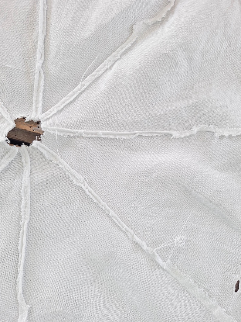 Puede incluir: Una falda de &aacute;rbol de tela blanca de forma circular con una abertura central y costuras radiales con bordes deshilachados. El tejido tiene una textura visible y est&aacute; construido con m&uacute;ltiples paneles unidos mediante costuras expuestas y sin rematar.