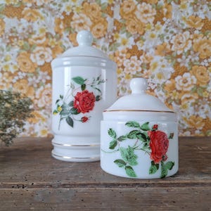 May include: Two white milk glass canisters with lids, featuring a red rose floral pattern with green leaves and thin gold trim accents. The taller canister is cylindrical, while the shorter canister is wider and more rounded in shape.