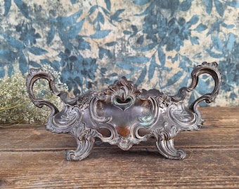 May include: An antique silver-toned centerpiece bowl with ornate scrollwork and handles. The bowl has a heart-shaped cutout and rests on four decorative feet. The background features a blue floral pattern.