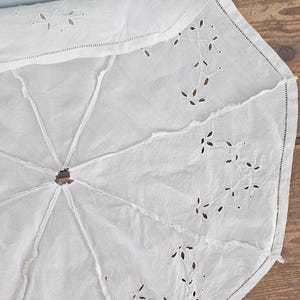 Puede incluir: Una funda de parasol de tela blanca con intrincados bordados de ojal en patrones de hojas. El tejido circular est&aacute; dividido en ocho paneles por costuras, con una abertura central y un dobladillo acabado a lo largo del borde exterior.