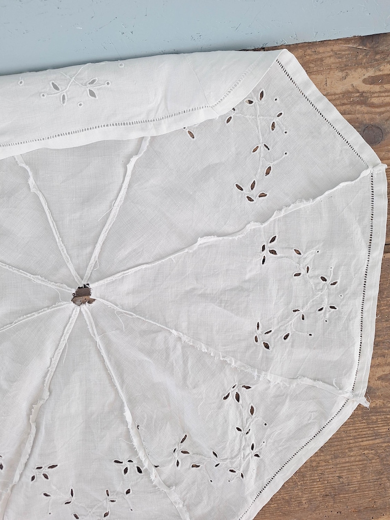 Puede incluir: Una funda de parasol de tela blanca con intrincados bordados de ojal en patrones de hojas. El tejido circular est&aacute; dividido en ocho paneles por costuras, con una abertura central y un dobladillo acabado a lo largo del borde exterior.