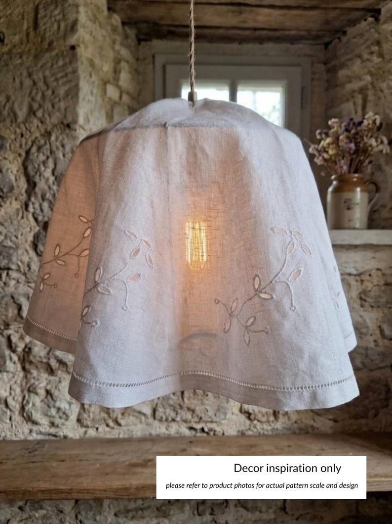 Puede incluir: Una pantalla de l&aacute;mpara colgante de tela blanca con delicados patrones de hojas bordadas y un dobladillo festoneado con costuras decorativas. La pantalla cuelga de un cord&oacute;n y contiene una bombilla de filamento brillante en el centro.