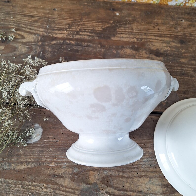 May include: A white ceramic soup tureen with a lid, featuring two side handles and a pedestal base. The tureen has a glossy finish and a slightly aged appearance, suggesting a vintage or antique style. The lid is partially visible.