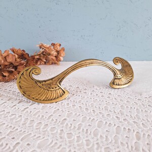 May include: A gold-toned metal drawer pull featuring a curved handle and two wide, fan-shaped ends with engraved linear detailing. The hardware has a weathered finish and a symmetrical design with scroll-like accents at the outer edges.