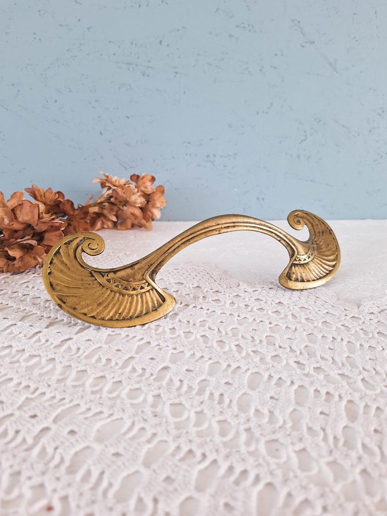 May include: A gold-toned metal drawer pull featuring a curved handle and two wide, fan-shaped ends with engraved linear detailing. The hardware has a weathered finish and a symmetrical design with scroll-like accents at the outer edges.