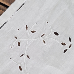 Puede incluir: Un tejido de lino blanco con delicados bordados en hilo blanco que forman un patr&oacute;n bot&aacute;nico de hojas y enredaderas. La tela presenta intrincados detalles de calados y un borde decorativo con vainica, mostrando una fina labor de costura.