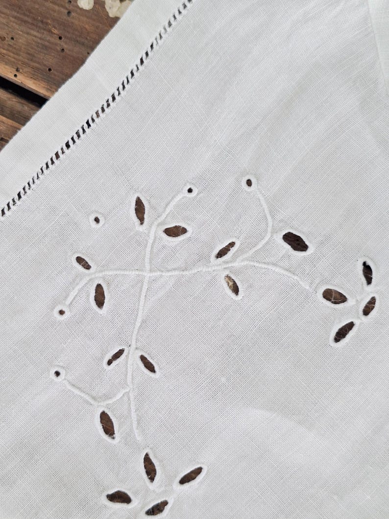 Puede incluir: Un tejido de lino blanco con delicados bordados en hilo blanco que forman un patr&oacute;n bot&aacute;nico de hojas y enredaderas. La tela presenta intrincados detalles de calados y un borde decorativo con vainica, mostrando una fina labor de costura.