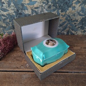 Puede incluir: Un joyero rectangular de cerámica turquesa con tapa redondeada que presenta un retrato circular de una persona con ropa de época. La caja está colocada dentro de un estuche gris texturizado con bisagras y forrado con tela dorada.
